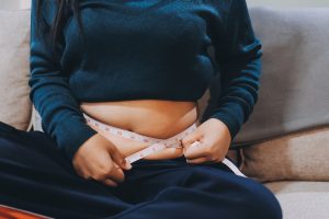 donna seduta sul divano , maglia alzata e si sta misurando con il metro il grasso addominale. ha la pancia gonfia