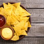 su tavolo di legno un piatto tondo pieno di nachos e due ciotole con salsa rossa e gialla