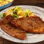 Paillard di vitello alla griglia: leggera, veloce e saporita 1 Su un tavolo di legno in un piatto fondo ci sono due paillard di vitello accanto ai cavoletti di Bruxelles.