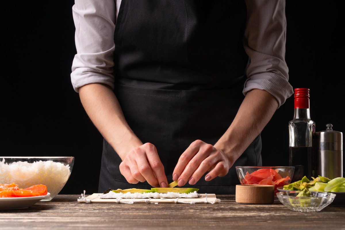 mani di una donna che cucina ricette giapponesi e sta per arrotolare sushi cin riso e avocado. Accanto sul tavolo ingredienti riso, avocado, salsa di sois e salmone