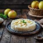 In primo piano su un tavolo di legno c'è un piatto fondo scuro con un risotto alle mele decorato con un fogliolina di menta. Sullo sfondo a sinistra c'è una ciotola con del riso e delle mele in un cestino. In primo piano sulla destra c'è un cucchiaio di legno.