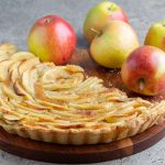 Torta di mele senza burro: video passo passo con foto 1 Su un tavolo di marmo grigio c'è un tagliere di legno con una torta di mele senza burro e sullo sfondo delle mele.