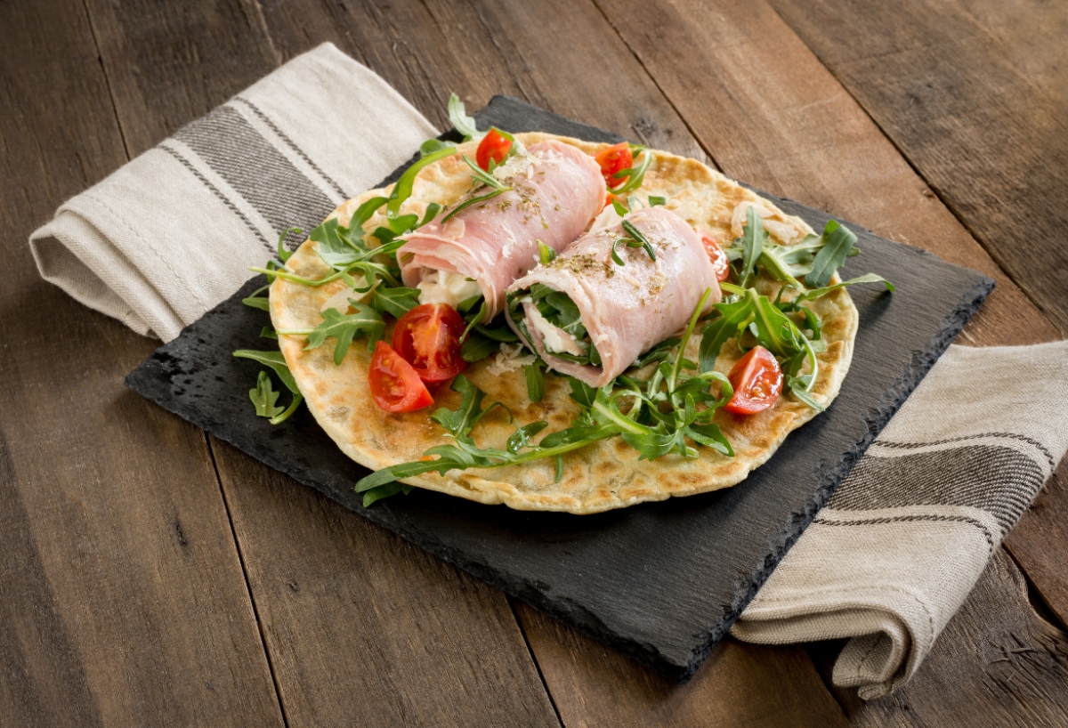su tavolo di legno, canovaccio a riga con tagliere di marmo nero e una focaccia con prosciutto e crescenza con sotto insalata e pomodorini