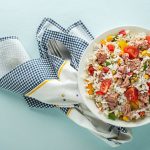 Insalata di riso con tonno: i consigli per farla perfetta 1 su sfondo azzurro, canovaccio a righe blu con una ciotola piena di insaltat di riso con tonno, pomodorini e e mais