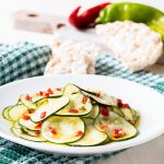 Zucchine marinate fatte in casa: ricetta facile e veloce 1 su canovaccio verde a righe bianche, un piatto con zucchine marinate, dietro gallette di riso e dietro peperoncini verdi e rossi