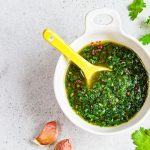 Chimichurri argentino: ricetta facile e veloce 1 Salsa chimichurri in una ciotola bianca con cucchiaio giallo, spicchi d’aglio e foglie di prezzemolo su un piano chiaro.