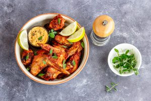 Iscriviti subito alla dieta Melarossa e scarica l'app 10 Alette di pollo in friggitrice ad aria servite in una ciotola con spicchi di limone, salsa speziata, erbe fresche e pepe su fondo grigio.