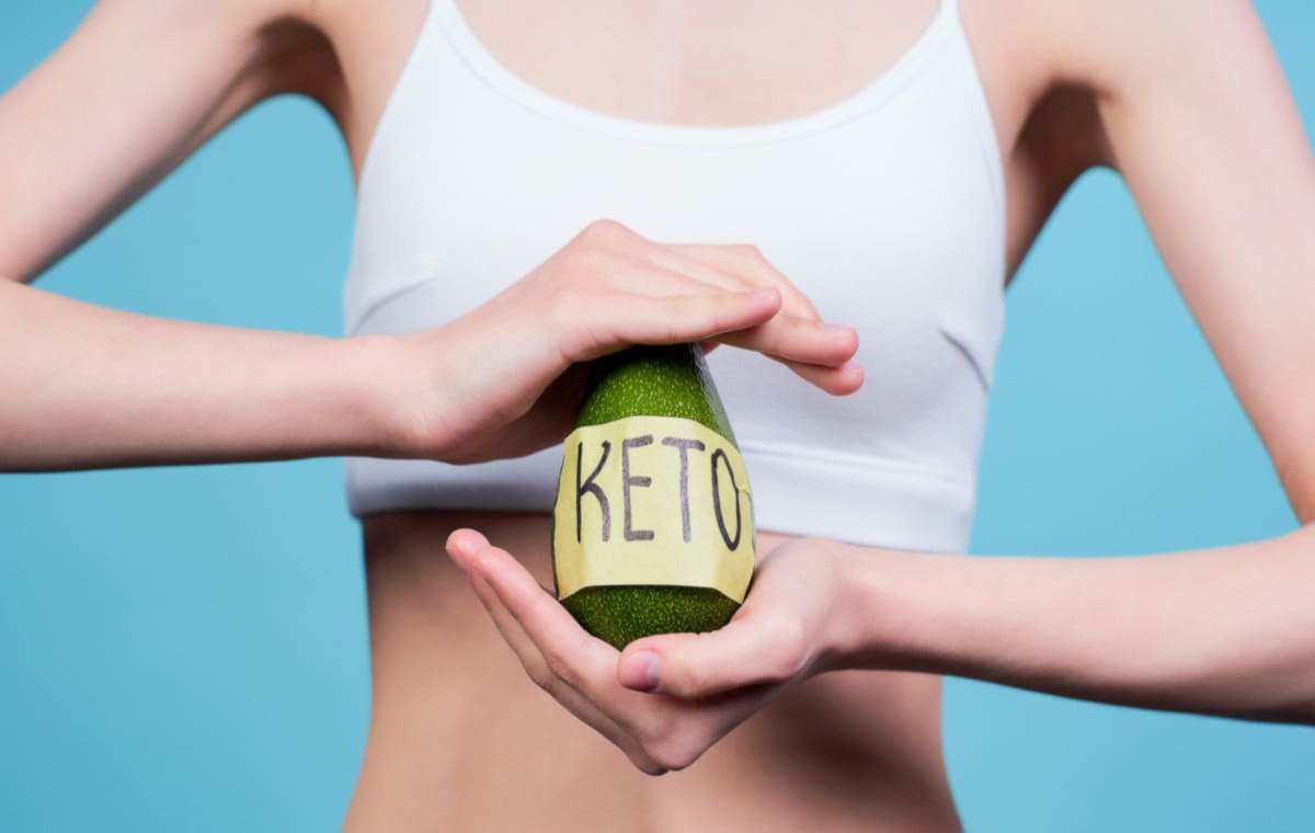 su sfondo blu donna con top bianco tiene in mano un avocado con scritta Keto perché segue la dieta chetogenica menu