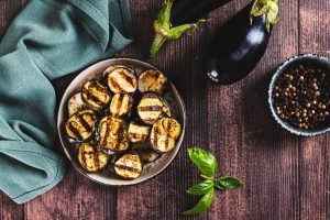 Iscriviti subito alla dieta Melarossa e scarica l'app 13 su tavolo di legno ciotola con melanzane in friggitrice ad aria. Accanto tovagliolo verde e sulla destra 2 melanzane e in un altra ciotola pepe nero