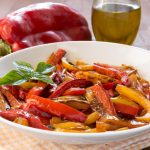 Peperoni in friggitrice ad aria: ricetta facile, veloce e leggera 1 su tavolo piatto bianco sotto un canovaccio giallo e bianco con peperoni in friggitrice ad aria. Accanto una bottiglia di olio EVO e un peperone rosso intero