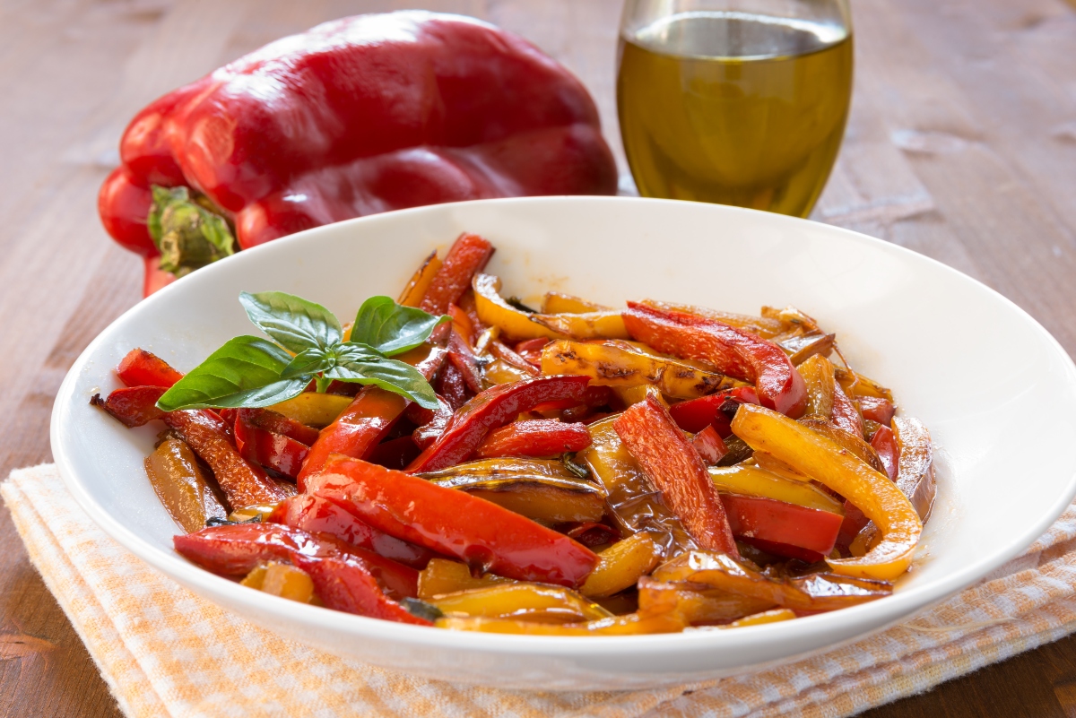 su tavolo piatto bianco sotto un canovaccio giallo e bianco con peperoni in friggitrice ad aria. Accanto una bottiglia di olio EVO e un peperone rosso intero