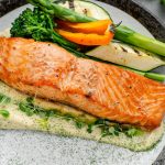 Salmone in friggitrice ad aria: ricetta facile e veloce 1 su tavolo da cucina primo piano di un piatto grigio con filetto di salmone in friggitrice ad aria con salsa allo yogurt e accanto verdure come asparagi, zucchine.