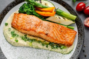 Iscriviti subito alla dieta Melarossa e scarica l'app 11 su tavolo da cucina primo piano di un piatto grigio con filetto di salmone in friggitrice ad aria con salsa allo yogurt e accanto verdure come asparagi, zucchine.