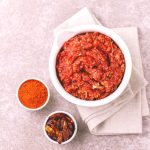 Salsa harissa fatta in casa: ricetta facile e speziata 1 In primo piano su uno sfondo di marmo chiaro c'รจ una ciotola bianca con della salsa harissa fatta in casa e accanto due ciotoline con peperoncini secchi e polvere di peperoncino.