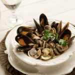 In primo piano su un piatto bianco in ceramica c'è un sauté di cozze e vongole con delle foglie di prezzemolo. Sullo sfondo un bicchiere di vino bianco.