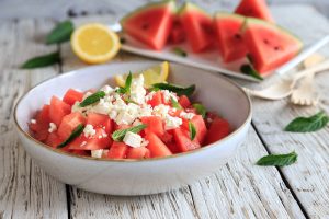 Iscriviti subito alla dieta Melarossa e scarica l'app 15 su tavolo di legno bianco ciotola con anguria e feta e foglie di menta; dietro un piatto bianco con anguria tagliata, mezzo limone foglie di menta e 2 forchette bianche