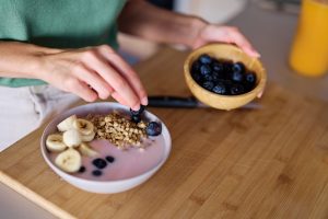 Iscriviti subito alla dieta Melarossa e scarica l'app 14 piano dall'alto di un tagliere in cucina con una donna che sta preparando la colazione con yogurt, cereali e banane: nell'altra mano ha una ciotola di mirtillo e si sta chiedendo se la frutta fa ingrassare.