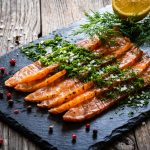 Gravlax: ricetta originale del salmone marinato svedese 1 su tavolo di legno tagliere di marmo con il gravlax di salmone. Sopra aneto e erbe aromatiche. attorno al tagliere pepe rosa e sale grosso