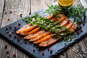Iscriviti subito alla dieta Melarossa e scarica l'app 14 su tavolo di legno tagliere di marmo con il gravlax di salmone. Sopra aneto e erbe aromatiche. attorno al tagliere pepe rosa e sale grosso