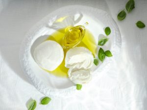 Iscriviti subito alla dieta Melarossa e scarica l'app 10 su tavolo con tovaglia bianca piatto bianco con mozzarella di bufala intera e una tagliata con olio EVO sotto e anche contenitore di olio attorno al piatto bianco foglie sparse di basilico