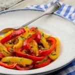 Peperoni in padella: contorno facile e leggero 1 In primo piano in un piatto fondo bianco con bordo azzurro ci sono dei peperoni in padella rossi e gialli e una forchetta. Sullo sfondo un tovagliolo a quadri bianchi e celesti.