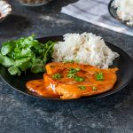 Pollo alla paprika: ricetta al forno facile e speziata 1 In primo piano piano su un piatto fondo scuro c'è una fettina di pollo alla paprika con del riso bianco e della verdura. Sullo sfondo c'è della paprika e un tovagliolo a quadri.