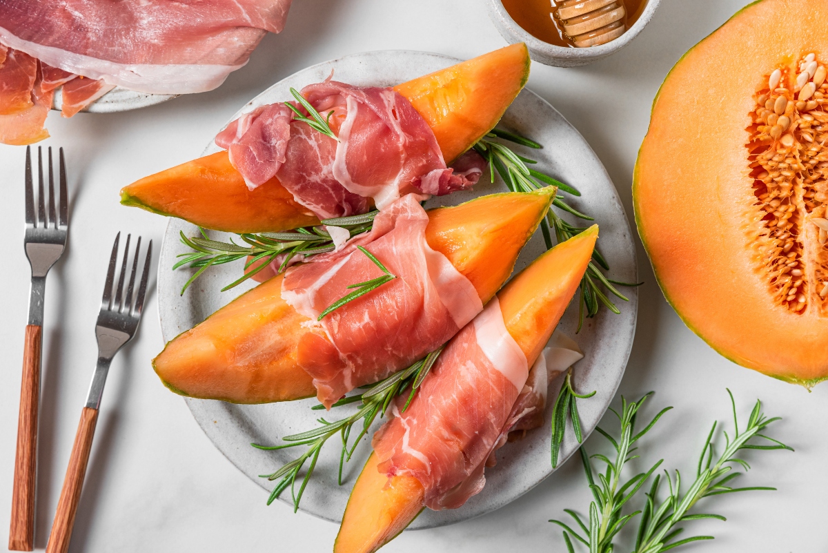 su tavolo bianco, piatto grigio con prosciutto e melone, sopra erbe aromatiche