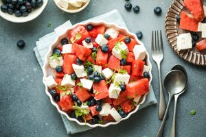 Iscriviti subito alla dieta Melarossa e scarica l'app 11 Ciotola di insalata con cubetti di anguria e feta, mirtilli ed erbe aromatiche, su uno sfondo grigio.