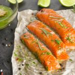 Salmone marinato: la ricetta fresca per un antipasto fresco e saporito 1 su tavolo di marmo carta da cucina dove sono sistemate 3 fette di salmone marinato. Sulla sinistra contenitore con la marinatura e un pennello, dietro fette di limone e erbe aromatiche. Attorno pepe bianco e rosa