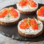 Cheesecake al salmone: l’antipasto cremoso con base croccante 1 In primo piano su un piatto tondo in ardesia ci sono delle mini porzioni di cheesecake al salmone decorate con dell'erba cipollina.