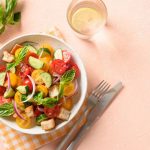 Cialledda materana: la ricetta fredda facile e gustosa 1 In primo piano su uno sfondo rosa chiaro c'è un piatto di cialledda materana fredda. Sullo sfondo un bicchiere di acqua con una fettina di limone, delle posate e un tovagliolo a quadri gialli e bianchi.