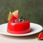 Gelo di melone: ricetta siciliana facile e leggera 1 In primo piano su un piatto fondo bianco, c'è il gelo di melone decorato con delle gocce di cioccolato, una fogliolina di menta e due spicchi di anguria. In primo piano sulla destra c'è un cucchiaino dorato con il gelo di melone.