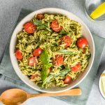 Pasta fredda al pesto: ricetta facile e veloce 1 In primo piano su uno sfondo chiaro e un canovaccio salvia, c'รจ un piatto di pasta fredda al pesto con pomodorini.
