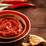 Salsa harissa fatta in casa: ricetta facile e speziata 1 In primo piano su un tavolo di legno scuro c'è una ciotolina color senape con della salsa harissa. Sullo sfondo un tovagliolo e dei peperoncini freschi.