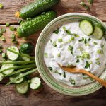 Salsa raita: la ricetta indiana fresca e speziata con yogurt 1 In primo piano su un tavolo di legno c'รจ una ciotola con della salsa raita ed erba cipollina. Sullo sfondo ci sono due piccoli cetrioli interi e alcuni tagliati a rondelle.