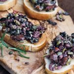 Salsa tapenade: la ricetta facile e veloce con olive nere 1 Crostini con salsa tapenade di olive nere e verdi e capperi, disposti su un tagliere di legno.