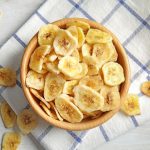 Chips di banana fatte in casa, sottili e croccanti 1 In primo piano su un canovaccio a righe bianco e blu, c'รจ una ciotola di legno con delle chips di banana croccanti.