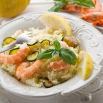 Cous cous con gamberi e zucchine: la ricetta semplice e gustosa 1 su tavolo, piatto bianco con cous con gamberi e zucchine con fette di limone e una forchetta