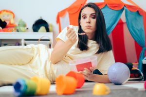 Iscriviti subito alla dieta Melarossa e scarica l'app 10 una giovane mamma distesa a terra nella camera dei suoi bambini con tutti i giocatoli a terra. Guarda il vuoto e sta mangiando una vaschetta di gelato perché ha un attacco di fame nervosa