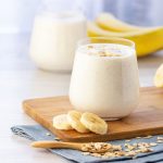 Frullato di banana: ricetta facile e varianti 1 In primo piano su un tagliere di legno c'รจ un bicchiere di vetro con il frullato di banana. Sullo sfondo ci sono delle banane fresche e un cucchiaino con dei fiocchi di avena.