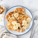 Pancake alla banana con farina d’avena: facili e veloci 1 In primo piano su un tavolo di marmo chiaro c'è un piatto con dei pancake alla banana decorati con qualche lamella di mandorla. Sullo sfondo una ciotolina con delle frutta secca.
