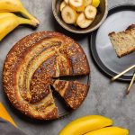 Torta di banane e yogurt: ricetta soffice senza burro 1 In primo piano su uno sfondo scuro c'รจ un torta di banane. Sulla destra un piattino nero con una fetta di torta e due cucchiaini. Intorno ci sono delle banane fresche.