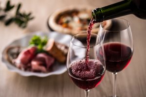 Iscriviti subito alla dieta Melarossa e scarica l'app 12 su tavolo di legno primo piano di una mano che serve il vino rosso con una mano in 2 calici. Dietro foto sfocata di 2 piatti di carne