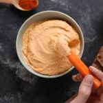 Hummus di carote fatto in casa, cremoso e delicato 1 su base grigio scuro ciotola vista dall'alto piena di humus di carote, due dita tengono una carota e prendono un pò di hummus
